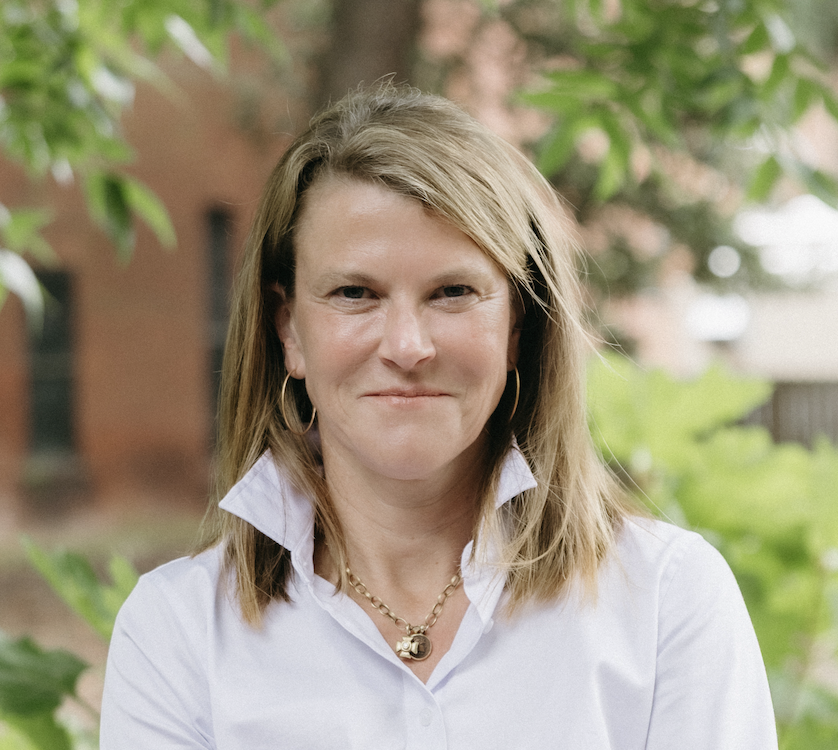 Laura smiling at the camera wearing a white shirt with arms crossed, greenery behind her.